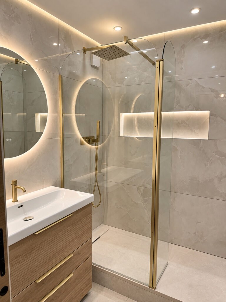 Salle de bain avec douche vitrée, robinetterie dorée, meuble en bois et miroirs rétroéclairés