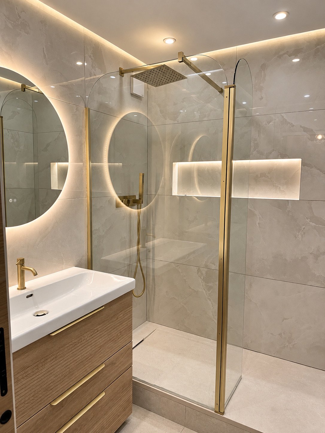 Salle de bain avec douche vitrée, robinetterie dorée, meuble en bois et miroirs rétroéclairés